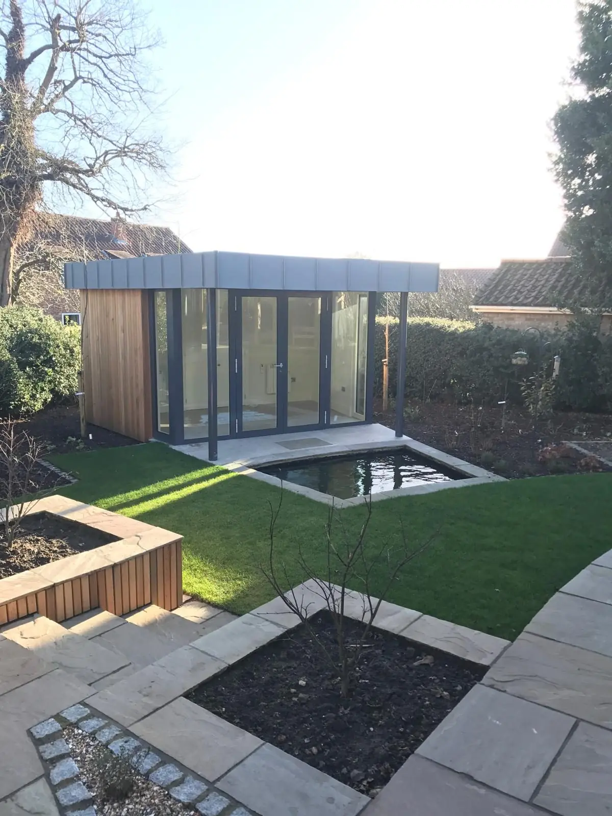 Cedar-clad garden room in Easingwold with a grey standing-seam roof and glazed folding doors, facing a wildlife pond