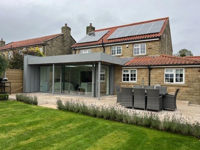 Single-storey extension to a stone house in Terrington, with full-height sliding glazing under a slim grey metal-clad roof